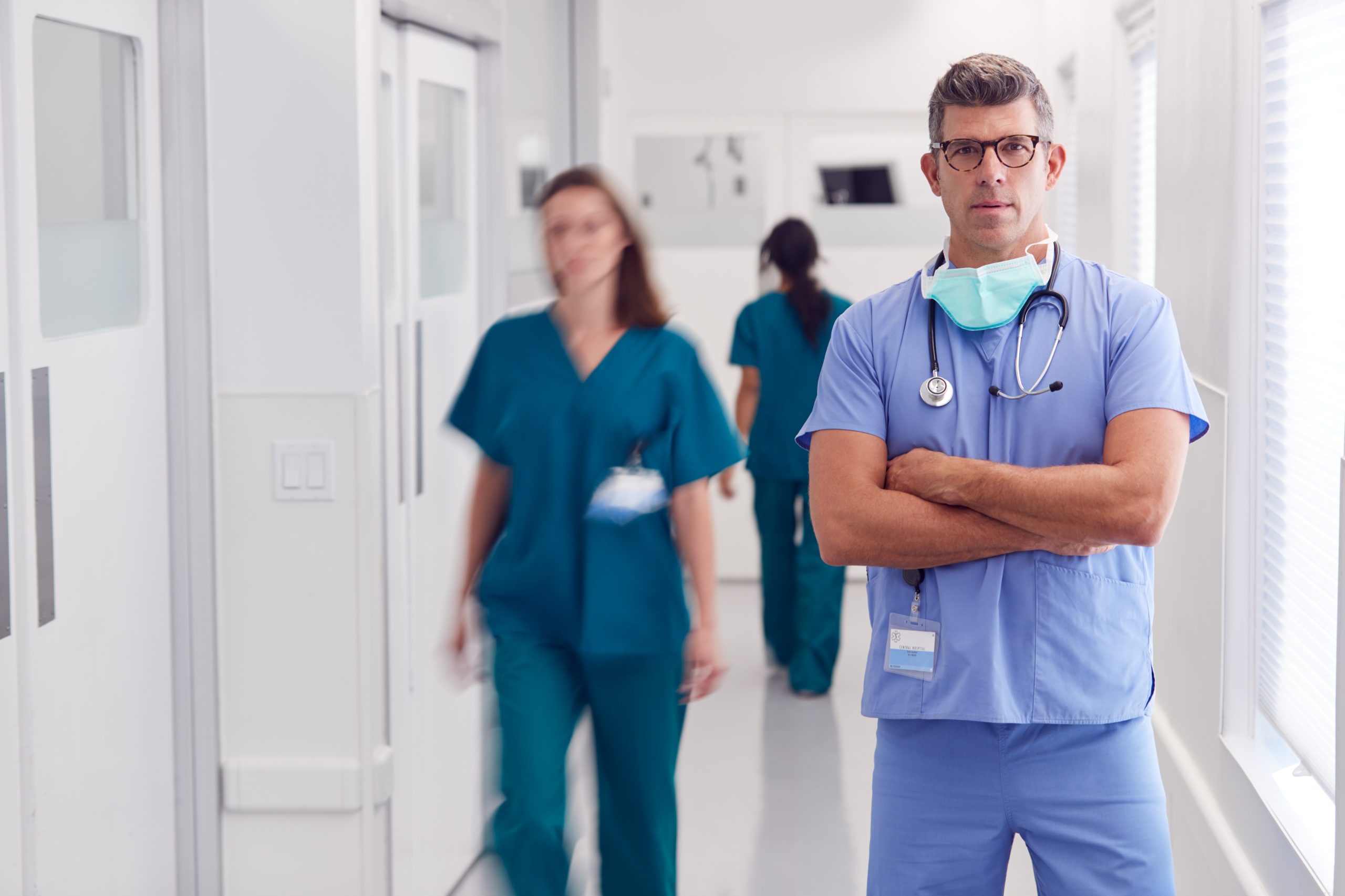 Portrait Of Mature Male Doctor Wearing Scrubs Standing In Busy Hospital Corridor